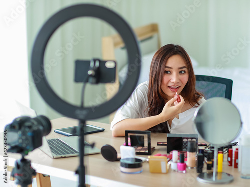 Selective focus on camera  Young and beautiful Asian girl shows make-up how to use lipstick to camera with smile and happy during broadcast video recording about cosmetics content and review.