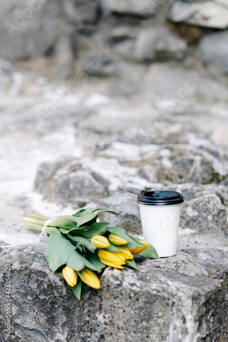 A bouquet of yellow tulips lies on a stone surface. Next to the bouquet is a paper take-away coffee cup.