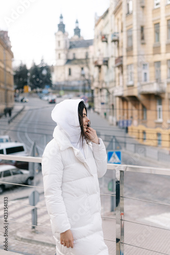 A girl in a white jacket and a hood walks along the sidewalk against the background of ancient architecture
