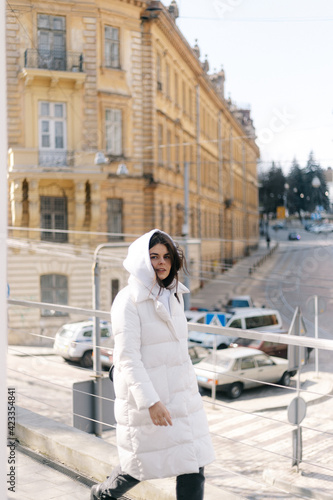 A girl in a white jacket and a hood walks along the sidewalk against the background of ancient architecture