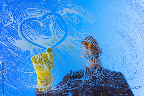 Fotografi Window cleaner or washer in yellow gloves drawing heart shape on a window glass