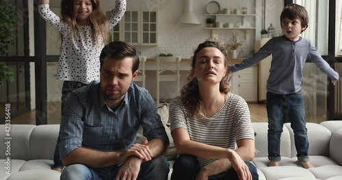 Tired couple sit on couch feels annoyed while noisy little daughter and son shouting jumping on sofa near parents. Too active hyperactive disobedient kids, need repose, upbringing difficulties concept