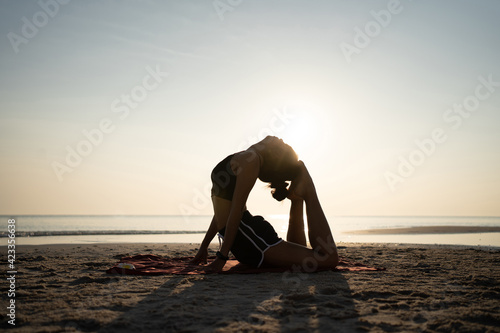 Silhouette of Asian woman practicing yoga at sunset. She arching her back and touching her head with her feet for a yoga pose at the beach. Health care concept.