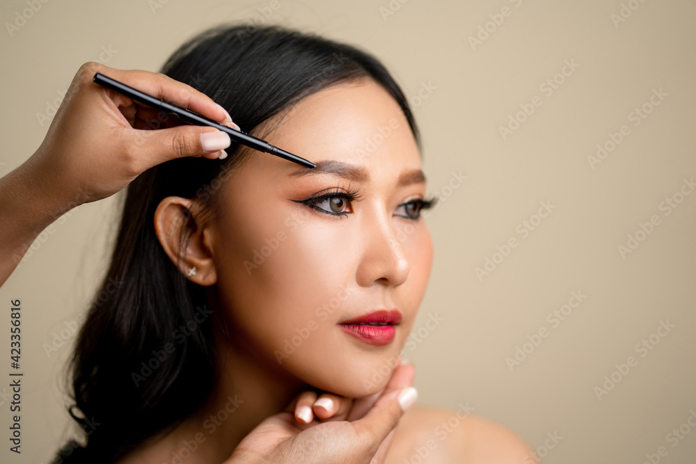 Makeup artist is drawing eyebrows on the face of a beautiful Asian woman.