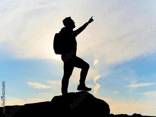Silhouette of a man with one arm raised and backpack. Pointing to the sky with one finger. Climber at the top of a mountain. Finished and celebrating the climb