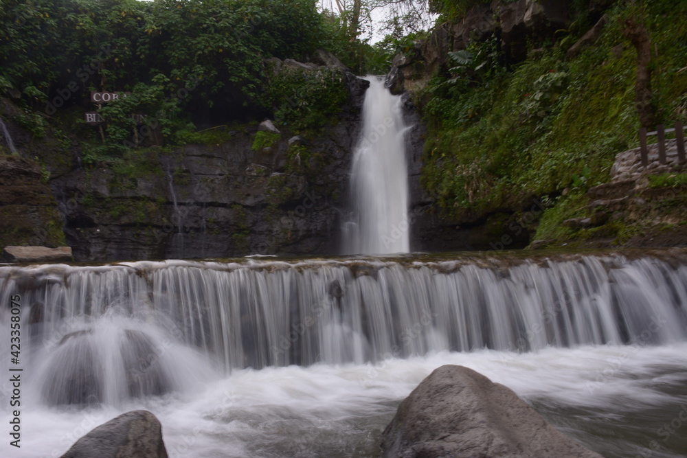 Fototapeta premium A stunning jungle waterfall flows into a cool mountain river with big rocks.