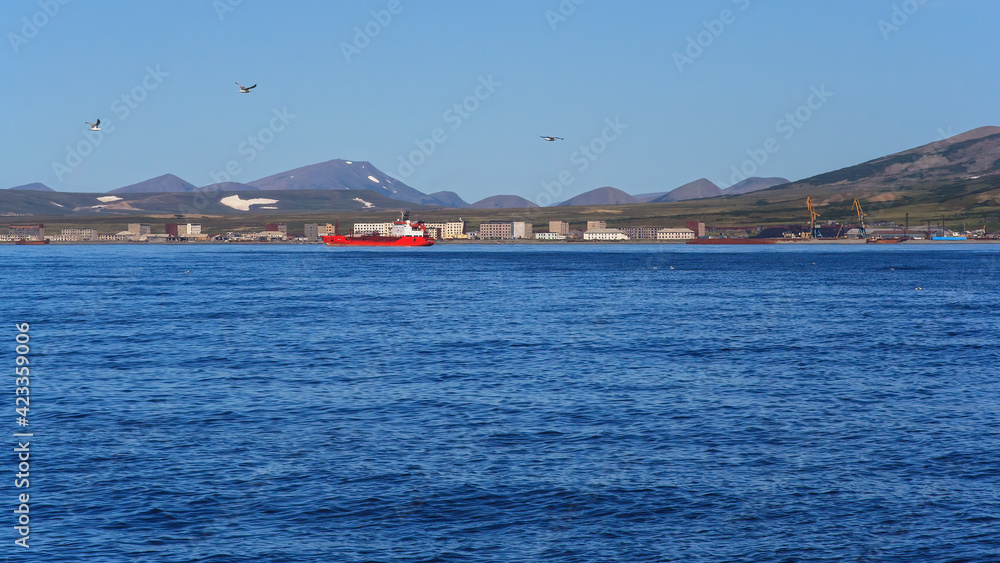 Summer seascape. A cargo ship near the coast. View of an abandoned northern settlement and an operating cargo seaport. In the distance tundra and mountains. Bering Sea. Beringovsky, Chukotka, Russia.