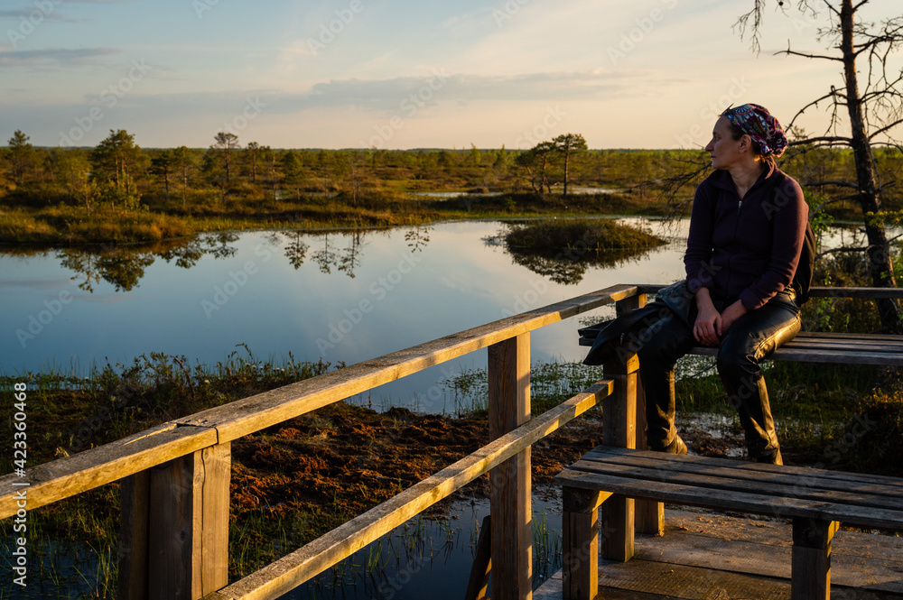 A woman admires the landscape of a forest lake among the raised bogs at sunset. A special observation platform for visitors to the reserve.