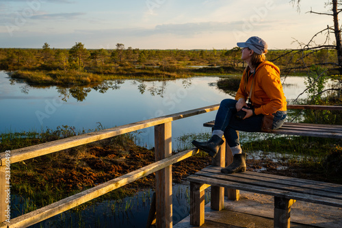 A female photographer admires the landscape of a forest lake among the raised bogs at sunset.