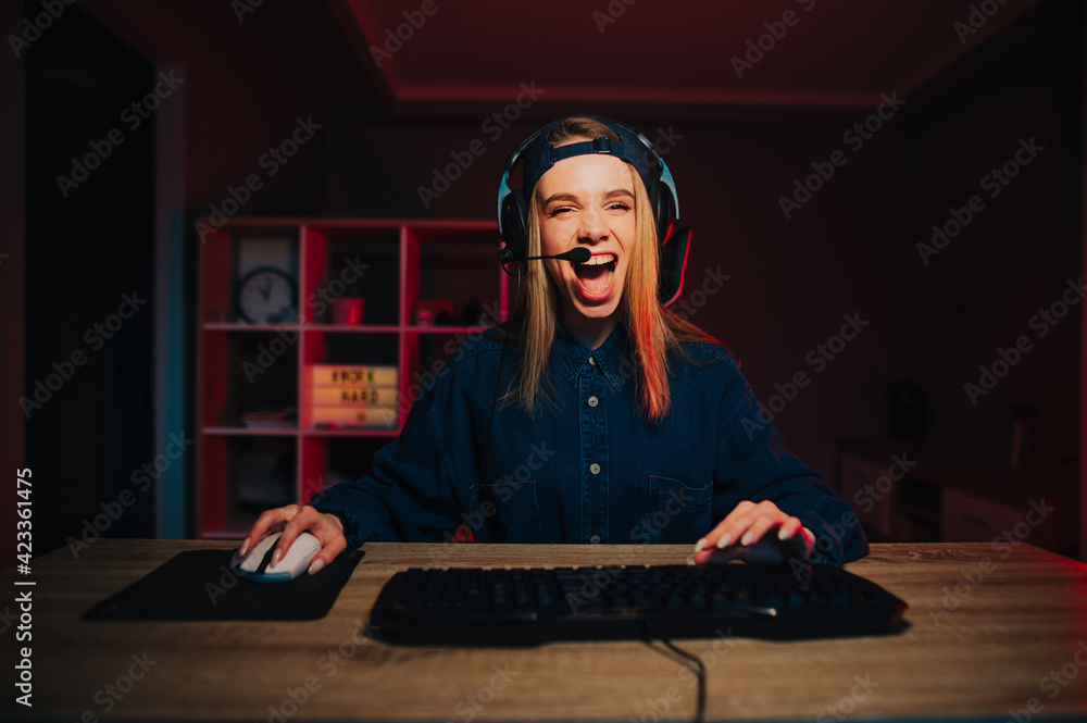 Joyful woman in a headset and a cap on her head plays and streams games with a happy emotional face, looks at the camera and shouts into the microphone.