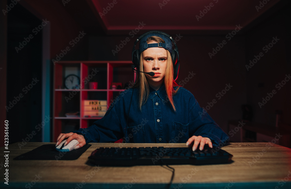 Displeased young gamer woman with headset on her head playing games at home on computer and frowning looking at camera on room background with red light. Displeased young gamer woman with headset on her head playing games at home on computer and frowning looking at camera on room background with red light.