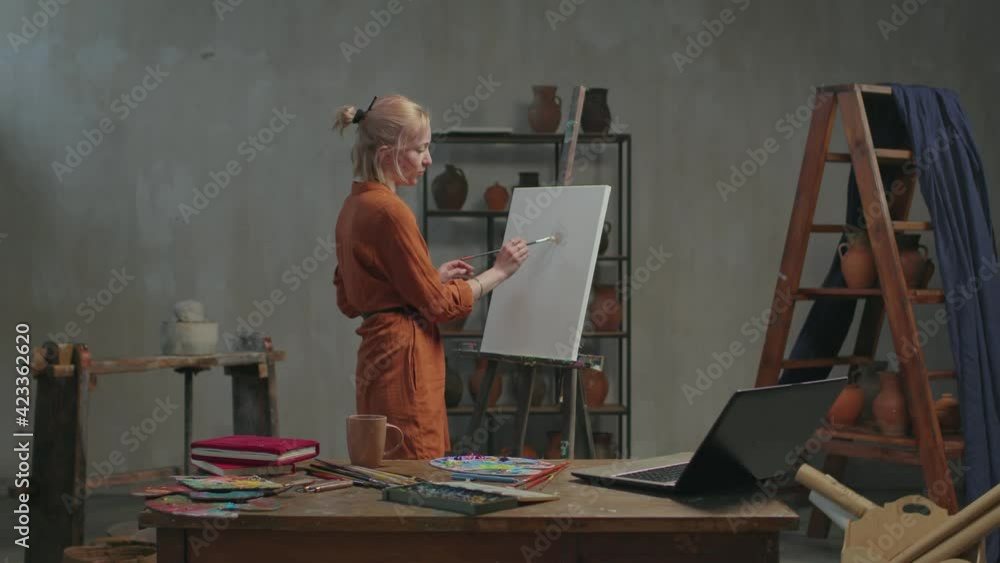 Professional female artist teaching art lesson online using laptop pc, live streaming tutorial video for vlog, explaining artistic techniques and skill of oil painting while working in art studio.