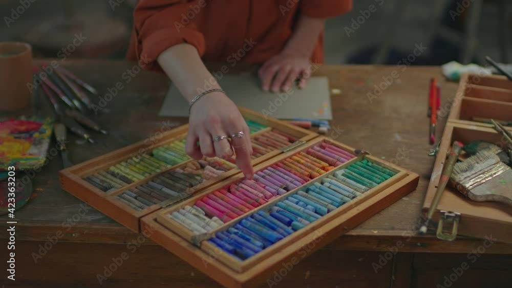 Close up of female artist hands selecting colors of pastel crayons from wooden set of multicolored chalks, with artistic tools placed on rustic table in art studio. Close up of female artist hands selecting colors of pastel crayons from wooden set of multicolored chalks, with artistic tools placed on rustic table in art studio.
