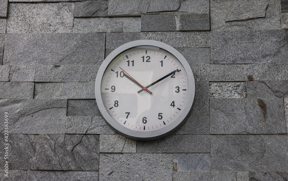 Modern wall clock on a wall tiled with rustic tiles. Modern white wall clock in sunlight with shadows on clock face on a grey background. Close up to a clock, with a red second pointer Modern wall clock on a wall tiled with rustic tiles. Modern white wall clock in sunlight with shadows on clock face on a grey background. Close up to a clock, with a red second pointer