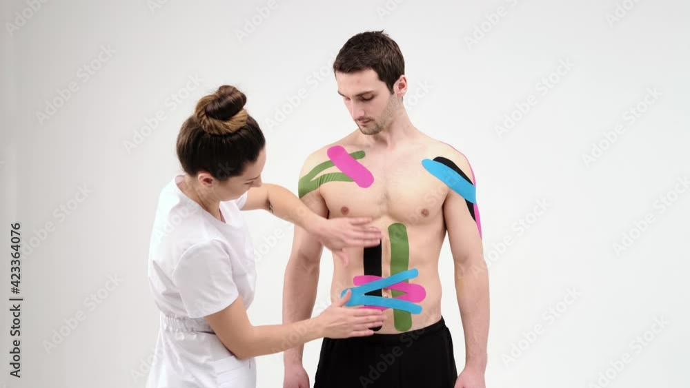 Female therapist applying kinesiology tape on a man's abdomen on the light background. Woman prepares male patient to glue kinesio adhesive tape on his belly. 4K Female therapist applying kinesiology tape on a man's abdomen on the light background. Woman prepares male patient to glue kinesio adhesive tape on his belly. 4K