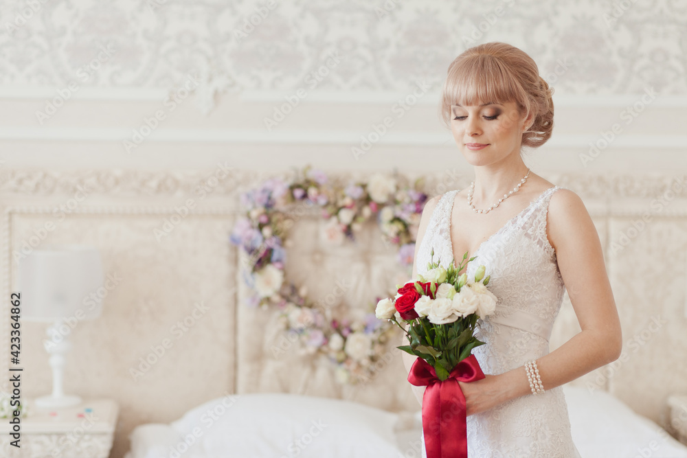 Pretty young Bride in elegant wedding dress with her bouquet. Blonde-haired woman with wedding hair-style in royal room of hotel. Boudoir morning of the bride in luxury interior. Wedding day ceremony Pretty young Bride in elegant wedding dress with her bouquet. Blonde-haired woman with wedding hair-style in royal room of hotel. Boudoir morning of the bride in luxury interior. Wedding day ceremony
