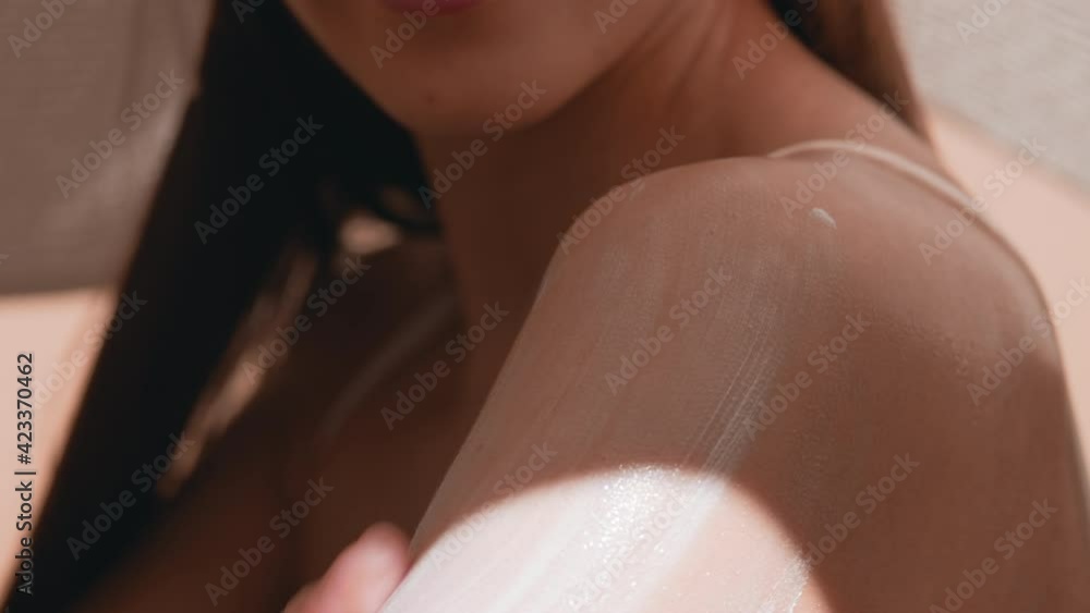 Young slim dark-haired woman in a big white hat and white swimwear puts sunscreen on her shoulder against beige background | Sunscreen putting on shot for skin care commercial Young slim dark-haired woman in a big white hat and white swimwear puts sunscreen on her shoulder against beige background | Sunscreen putting on shot for skin care commercial