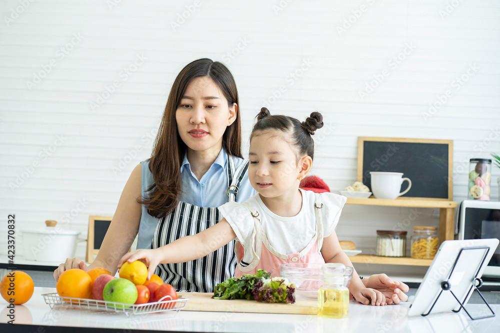 A cute daughter picks up an orange, prepares a salad, talks to the mother, and she takes a cooking class through the internet in the kitchen. 
