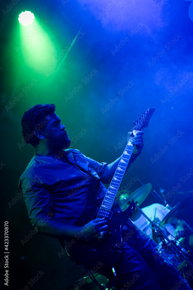 Obraz premium Musician playing electric guitar during rock band live concert. Black hair male guitar player performing on stage. Music foggy lights show