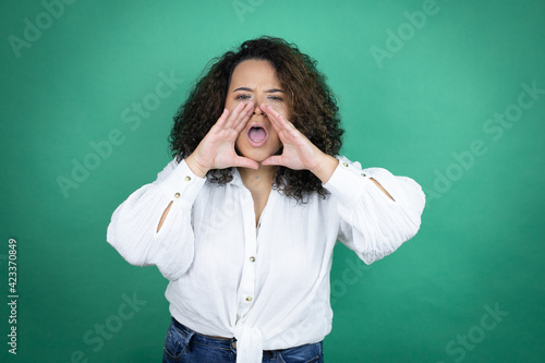 Young african american girl wearing white shirt over green background shouting and screaming loud to side with hands on mouth