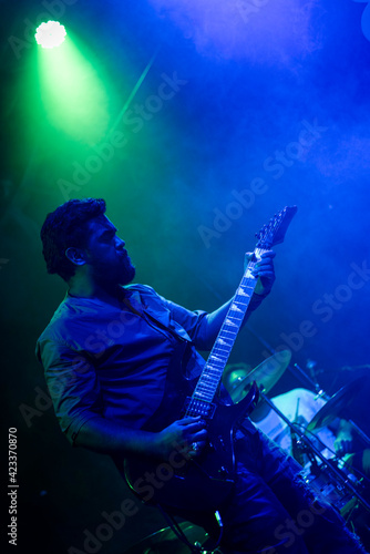 Musician playing electric guitar during rock band live concert. Black hair male guitar player performing on stage. Music foggy lights show