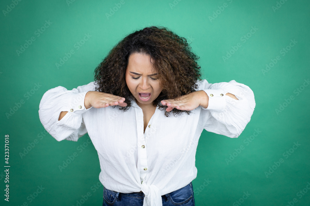 Young african american girl wearing white shirt over green background shouting and screaming loud down with hands on mouth Young african american girl wearing white shirt over green background shouting and screaming loud down with hands on mouth