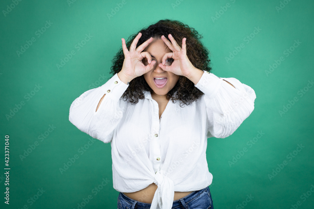Young african american girl wearing white shirt over green background doing ok gesture shocked with smiling face, eye looking through fingers