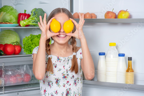 Smiling beautiful young teen girl holding yellow lemons over eyes while standing near open fridge in kitchen at home
