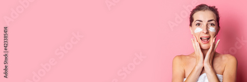 beautiful young woman applies cream to her face on a pink background. Banner.