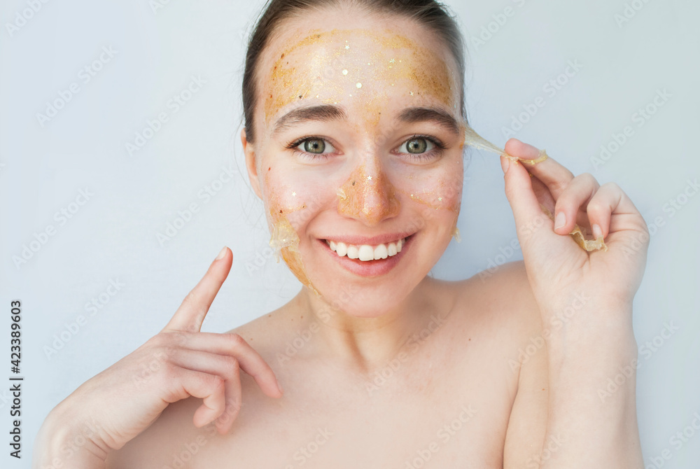 A beautiful girl takes off a yellow cosmetic mask from her face. Surprised face, shows a finger. Light background. A beautiful girl takes off a yellow cosmetic mask from her face. Surprised face, shows a finger. Light background.