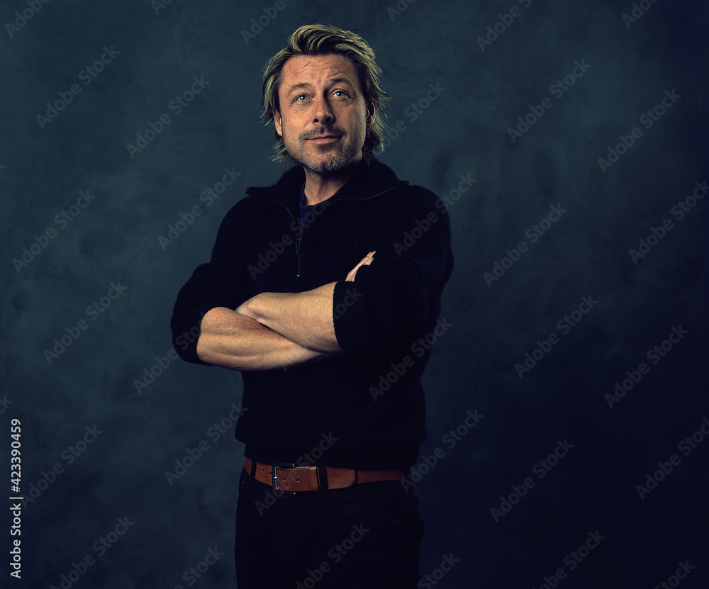 Relaxed blonde man with stubble beard in black wool sweater and jeans in front of a dark wall. Relaxed blonde man with stubble beard in black wool sweater and jeans in front of a dark wall.