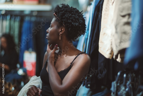 A beautiful young black woman with a hanger in hand is pensively looking around while standing in a clothing store with various textiles and clothes hanging around  shallow depth of field © skyNext