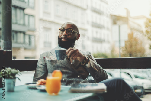 The portrait of a handsome stylish wealthy African guy with a beautiful black beard, in glasses, bald, in an elegant suit, sitting on a rainy morning in a street cafe and drinking delicious coffee photo