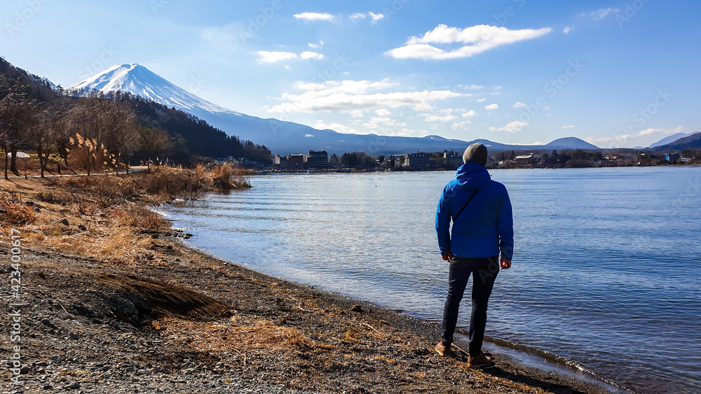 A man standing at the side of Kawaguchiko Lake, Japan with the view on Mt Fuji. The magnificent mountain surrounded by clouds. Soft reflections in the calm surface of the lake. Serenity and calmness