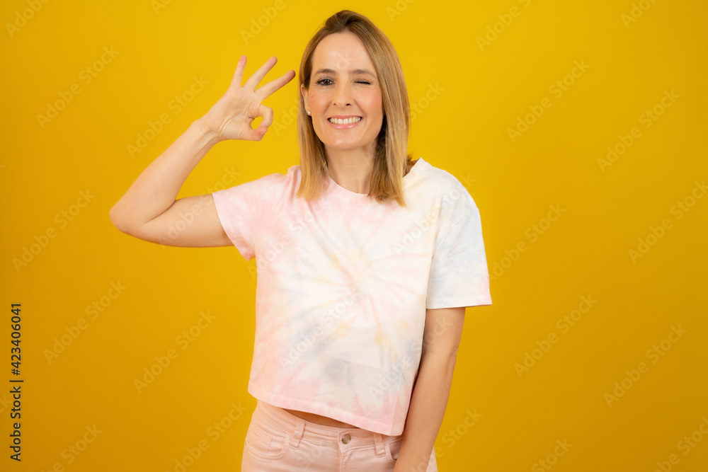 Young beautiful woman wearing summer clothes standing over yellow isolated background smiling positive doing okay sign with hand and fingers. Successful expression. Young beautiful woman wearing summer clothes standing over yellow isolated background smiling positive doing okay sign with hand and fingers. Successful expression.