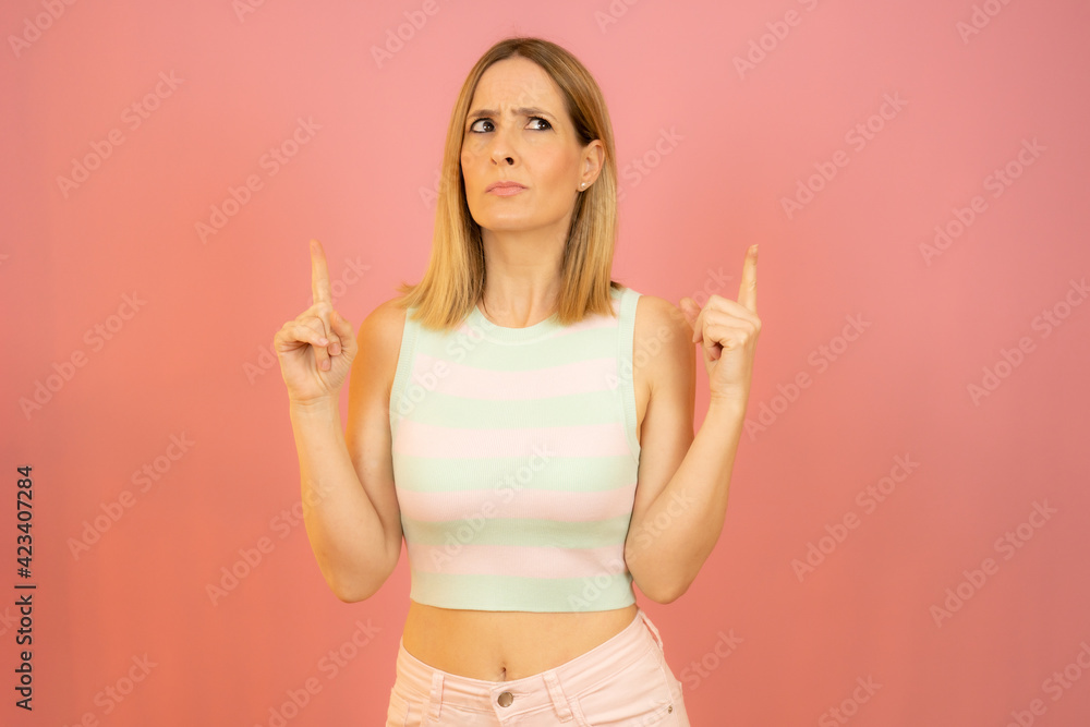Portrait of a young woman dressed in summer clothes pointing fingers up at copy space isolated over pink background