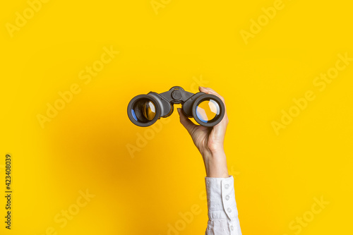 female hand holds black binoculars on a bright yellow background