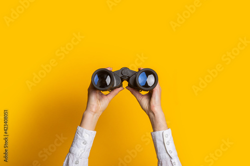 female hands hold black binoculars on a bright yellow background photo