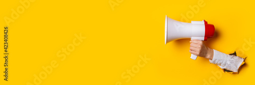 Fotografia Hand holds a megaphone from a hole in the wall on a yellow background