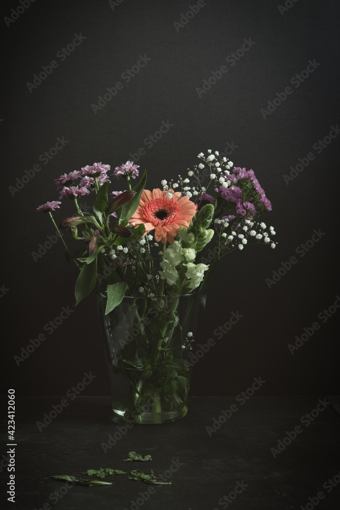 Fototapeta premium Vertical still life with a mix of spring flowers. An orange gerbera, red carnation, purple chrysanthemums and white gypsophila in a glass vase. Dark moody studio shot with copy space.