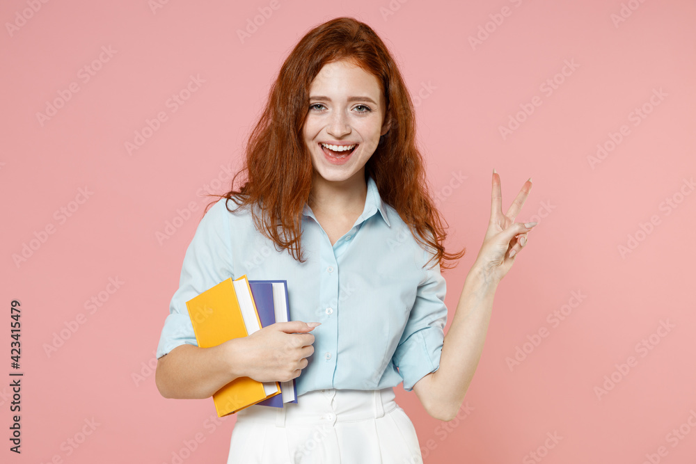 Young happy fun redhead student woman in blue shirt hold books notebook show victory v-sign gesture isolated on pastel pink background studio portrait Education high school university college concept Young happy fun redhead student woman in blue shirt hold books notebook show victory v-sign gesture isolated on pastel pink background studio portrait Education high school university college concept