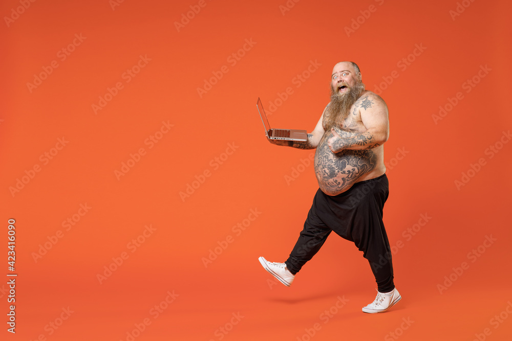 Full length side view fat fun pudge obese chubby overweight tattooed bearded big belly man in pants naked torso hold laptop pc computer walk show thumb up gesture isolated on orange background studio.