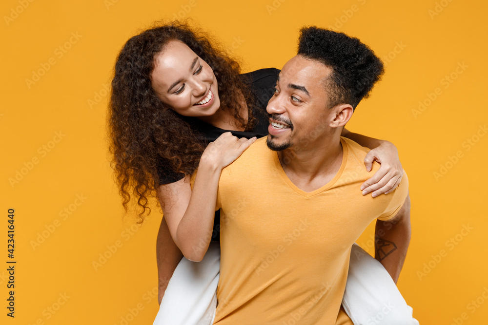 Young couple two together family smiling happy african man woman 20s wearing black t-shirt giving piggyback ride to joyful sit on back look to each other isolated on yellow background studio portrait. Young couple two together family smiling happy african man woman 20s wearing black t-shirt giving piggyback ride to joyful sit on back look to each other isolated on yellow background studio portrait.