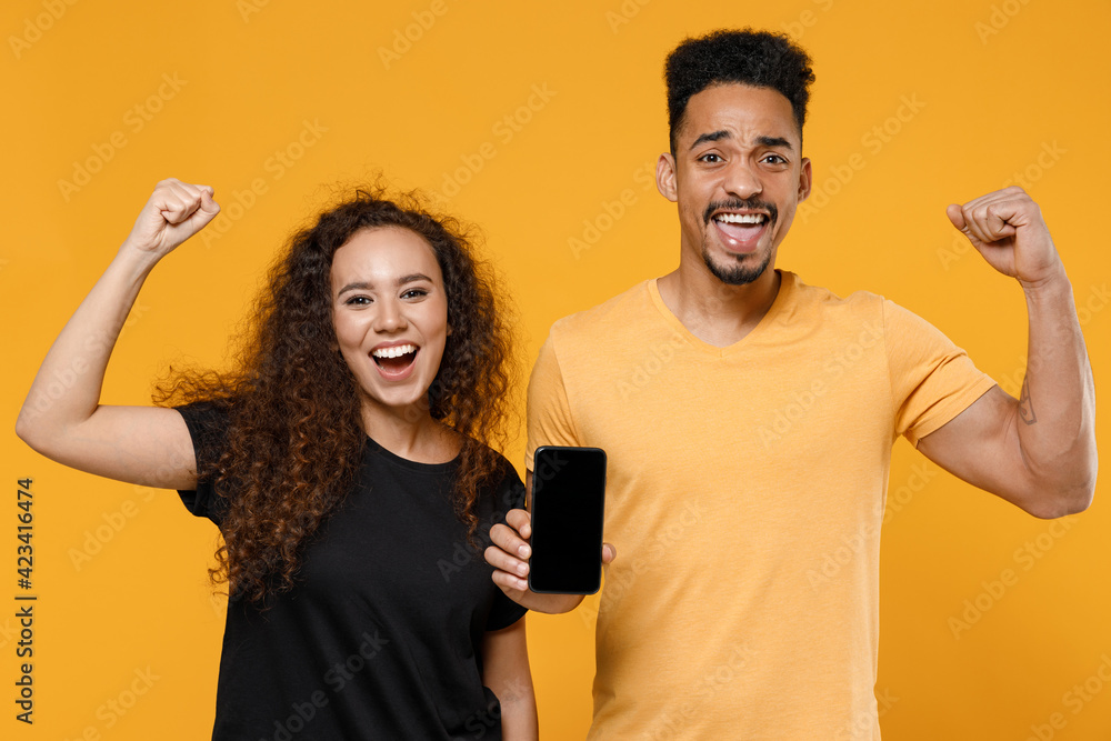 Young couple two friends together african man woman in black t-shirt hold mobile phone, blank screen workspace do winner gesture clench fist celebrate isolated on yellow background studio portrait. Young couple two friends together african man woman in black t-shirt hold mobile phone, blank screen workspace do winner gesture clench fist celebrate isolated on yellow background studio portrait.