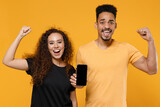 Young couple two friends together african man woman in black t-shirt hold mobile phone, blank screen workspace do winner gesture clench fist celebrate isolated on yellow background studio portrait.