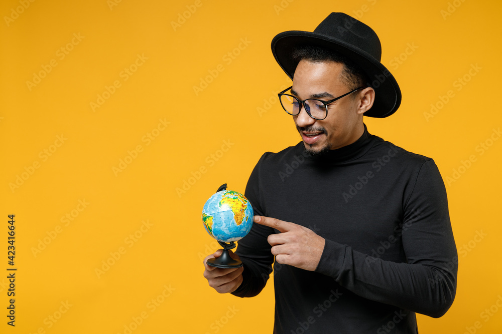 Fototapeta premium Young happy geographer smiling dreamful 20s wearing stylish black shirt hat eyeglasses holding in hands spin point index finger on world globe isolated on yellow orange background studio portrait