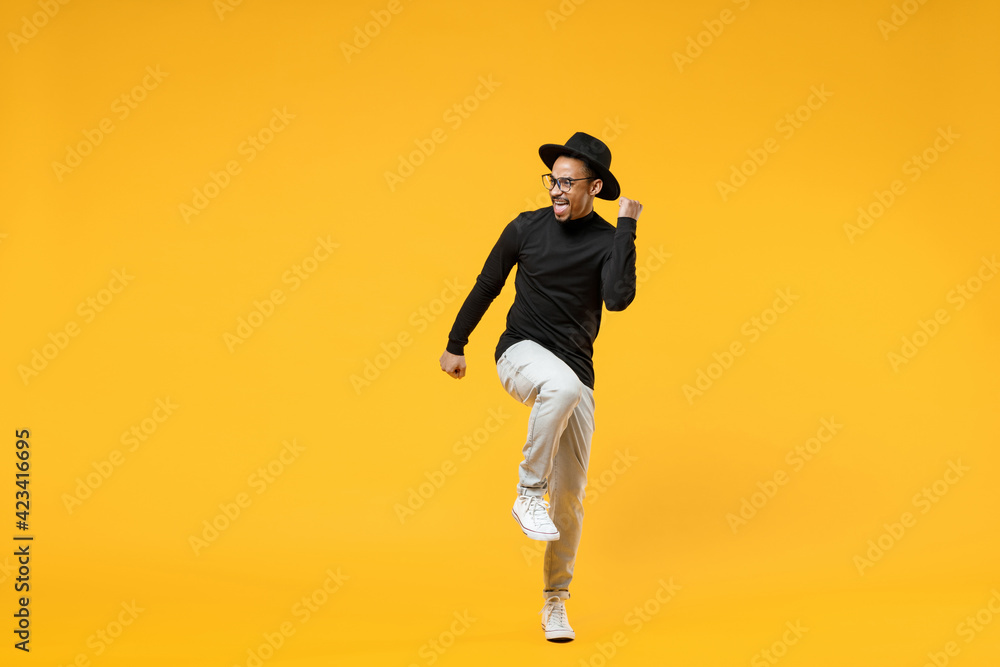 Fototapeta premium Full length young fun african man 20s wearing stylish black hat shirt eyeglasses do winner gesture clench fist celebrating ith raised up leg isolated on yellow orange color background studio portrait