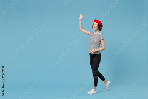 Full length of young smiling woman with short hairdo in french beret red hat striped t-shirt hold hands crossed folded walk going waving hand greet isolated on pastel blue background studio portrait.