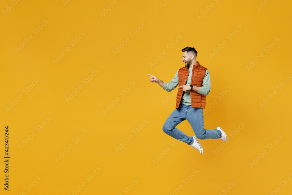 Full length side view young happy fun caucasian man 20s wear orange vest mint sweatshirt jump high point index finger aside on copy space workspace mock up area isolated on yellow background studio.