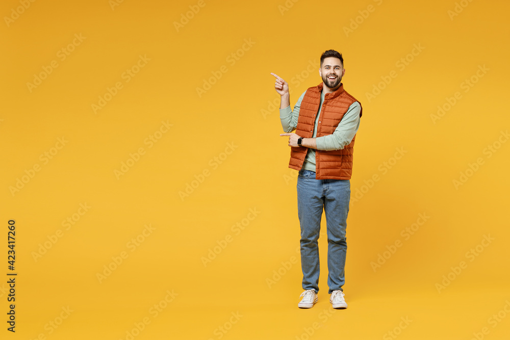 Full length young smiling happy confident smiling cheerful fun caucasian man in orange vest mint sweatshirt point index finger aside on copy space area mock up isolated on yellow background studio Full length young smiling happy confident smiling cheerful fun caucasian man in orange vest mint sweatshirt point index finger aside on copy space area mock up isolated on yellow background studio